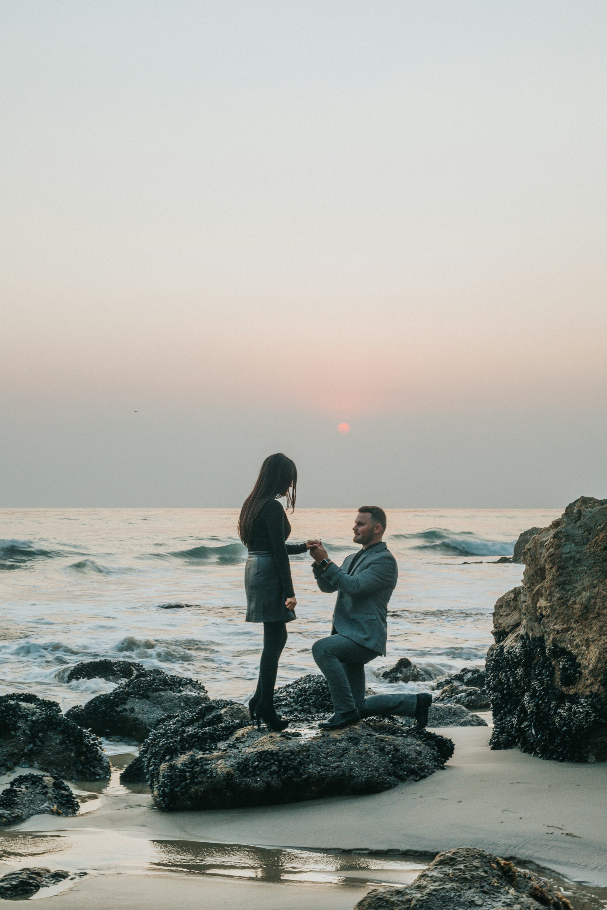 PLACEHOLDER (Unsplash) — man kneeling in front of woman on a rock at the beach